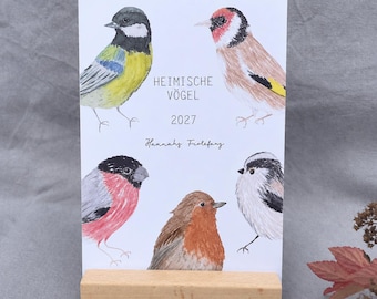 May include: A 2027 calendar card titled HEIMISCHE VÖGEL featuring watercolour illustrations of five birds, including a great tit, goldfinch, bullfinch, robin, and long-tailed tit, displayed on a rectangular light-coloured wooden block stand.