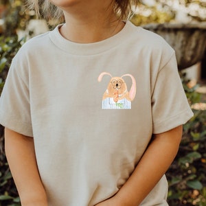 May include: A child wearing a light beige t-shirt with a cartoon bunny wearing glasses and a blue and white striped shirt printed on the front. The child is smiling and standing in front of a green bush.