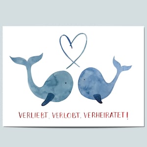 May include: A watercolor illustration features two blue whales facing each other beneath a blue heart. The whales have dark blue accents. The text "VERLIEBT, VERLOBT, VERHEIRATET!" is written in red below the whales.