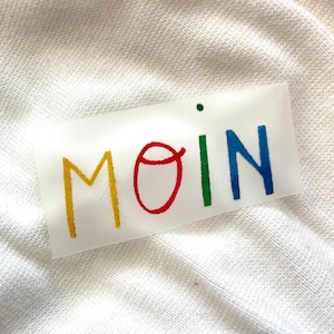 Peut inclure: Un autocollant transparent avec le mot "MOIN" écrit en lettres colorées. Les lettres sont jaunes, rouges, vertes et bleues.