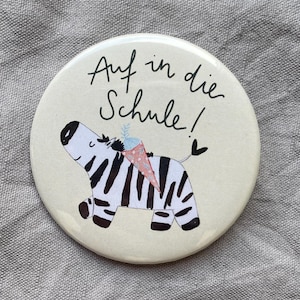 Könnte beinhalten: Ein runder, cremefarbener Ansteckbutton mit einer schwarz-weißen Illustration eines Zebras, das eine rosa, gepunktete Schultüte auf dem Rücken trägt. Der Text "Auf in die Schule!" ist in schwarzer Handschrift über dem Zebra gedruckt.