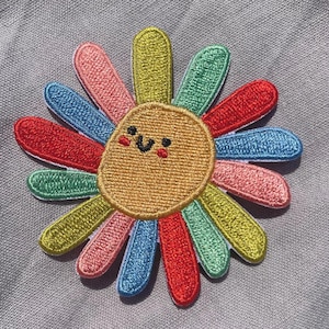 Op de afbeelding: Geborduurde patch met een lachende zonnebloem met kleurrijke bloemblaadjes in rood, roze, blauw, groen en geel. De patch heeft een lichtbruin midden en staat tegen een grijze achtergrond.