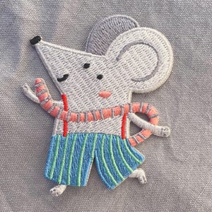 Peut inclure: Écusson brodé représentant une souris de dessin animé avec une tête et des oreilles grises, portant un short rayé bleu et une écharpe rose. La souris a des bretelles rouges et est en position de course. L'écusson est sur un fond de tissu gris.