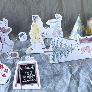 May include: A set of paper cutouts featuring a winter scene with a deer, a goose, a hedgehog, a snowman, a Christmas tree, a house, a person pulling a sled, a person carrying gifts, and a sign that reads "Weihnachts G&auml;nse bl&uuml;mchen".