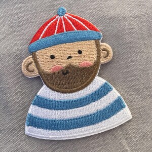 Könnte beinhalten: Gestickter Aufnäher mit einem Cartoon-Seemann. Der Aufnäher zeigt eine rot-blau gestreifte Mütze, einen braunen Bart und ein blau-weiß gestreiftes Oberteil. Das Design ist skurril, mit rosa Wangen, und eignet sich für Kleidung oder Accessoires.
