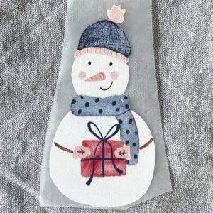 Pode incluir: Um boneco de neve branco com um chapéu e uma echarpe azul e branca segurando uma caixa de presente vermelha com um laço branco. O boneco de neve está sobre um fundo branco.