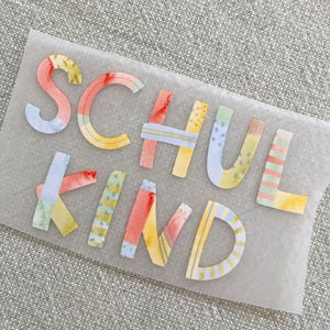 Könnte beinhalten: Ein farbenfroher, gemusterter Bügelaufkleber mit den Worten "Schulkind" in Deutsch, was "Schulkind" bedeutet.