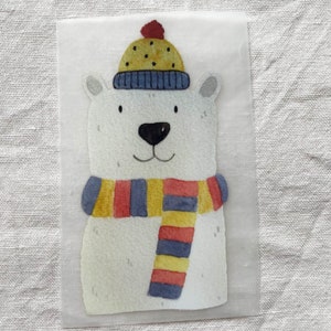 Könnte beinhalten: Ein weißer Eisbär, der einen blau-gelb gestreiften Schal und eine gelbe Mütze mit roten Punkten trägt. Der Bär befindet sich auf einem weißen Hintergrund.