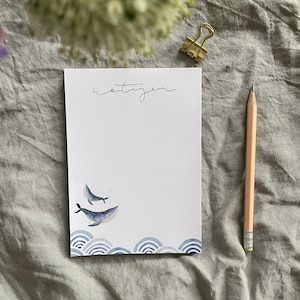 Könnte beinhalten: Ein weißer Notizblock mit dem Wort "notizen" in schwarzer Tinte oben. Der Notizblock hat eine blaue Aquarell-Illustration von zwei Walen, die im Meer schwimmen, mit Wellen am Boden.