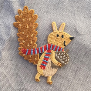 Peut inclure: Écusson brodé représentant un écureuil de dessin animé avec une queue touffue et une écharpe rayée. L'écureuil tient un gland. Les couleurs sont le beige, le marron, le rouge et le bleu.