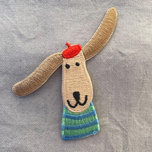 May include: Embroidered patch of a cartoon dog with long ears, wearing a red beret and a blue and green striped shirt. The patch is beige, with black eyes and a black nose. The dog's ears are brown.