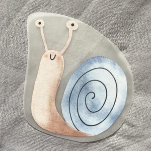 Könnte beinhalten: Ein Aufkleber mit einer niedlichen Schnecke mit hellrosa Körper und blau-weißem Spiralgehäuse. Die Schnecke hat zwei Fühler mit schwarzen Punkten und ein lächelndes Gesicht. Der Aufkleber befindet sich auf grauem Hintergrund.
