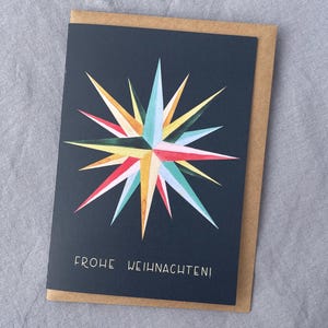 May include: A greeting card with a navy blue background and a colorful starburst design in red, yellow, turquoise, and white. The card reads "FROHE WEIHNACHTEN!" in white lettering and is inside a brown envelope.