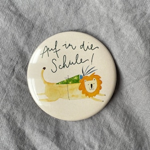 Pode incluir: Um botão redondo branco com um leão de desenho animado usando uma mochila verde e o texto "Auf in die Schule!" em preto.