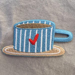 Może przedstawiać: Haftowana naszywka przedstawiająca niebieski kubek z białymi pionowymi paskami i czerwonym znakiem wyboru. Kubek stoi na beżowym spodku, z brązowym wnętrzem i niebieskim uchwytem. Do odzieży lub akcesoriów.