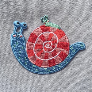 Könnte beinhalten: Ein gestickter Aufnäher, der eine blaue Schnecke mit einem rot-rosa karierten Spiralgehäuse zeigt. Die Schnecke hat zwei Augenstiele, ein lächelndes Gesicht mit rosa Wangen und kleine grüne Punkte auf dem Körper. Eine winzige grüne Schnecke sitzt auf dem Gehäuse.