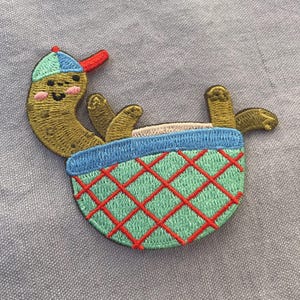 Può includere: Toppa ricamata con un cetriolo cartoon con un berretto da baseball, che si rilassa in un cestino. Il cetriolo è verde oliva con un viso sorridente e guance rosa. Il cestino è blu e verde con un motivo a rombi rossi.