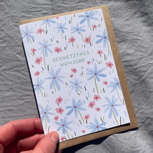 May include: A birthday greeting card with a floral design. The card features light blue flowers and pink flowers on a white background. The words "GEBURTSTAGS W&Uuml;NSCHE" are printed in green. The card is held in a person's hand.