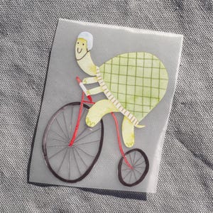 Op de afbeelding: Een grillige sticker met een gele schildpad op een rode fiets. De schildpad draagt een witte helm en heeft een groen geruit schild. De fiets heeft grijze wielen met rode spaken, tegen een grijze achtergrond.