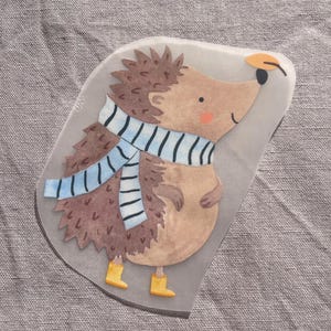 Könnte beinhalten: Eine skurrile Illustration eines Igels mit einem blau-weiß gestreiften Schal und gelben Stiefeln. Der Igel hat braunes Fell und ein hellbraunes Gesicht mit einem kleinen orangefarbenen Hut. Das Bild befindet sich auf einem transparenten, unregelmäßig geformten Hintergrund.