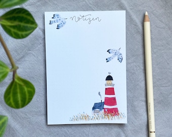 May include: A white notepad with a lighthouse illustration, two seagulls, and the word "notizen" in script. A white pencil rests beside the notepad. The lighthouse is red and white.