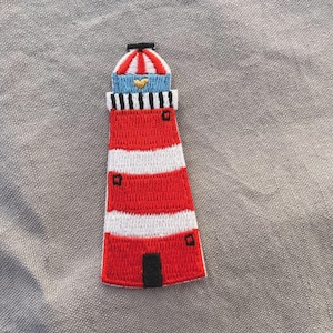 Op de afbeelding: Geborduurde patch van een vuurtoren met rood-witte strepen, een blauwe top met een gouden hart en een zwarte deur. De patch heeft een nautisch thema.