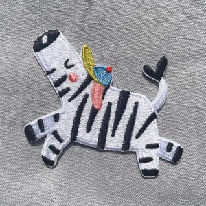 Può includere: Una toppa ricamata termoadesiva raffigurante una zebra bianca con strisce nere, una guancia rosa e un piccolo berretto giallo e blu sulla testa. La zebra è raffigurata in una posa giocosa con la coda alzata, mostrando cuciture dettagliate.