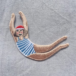 Può includere: Una toppa ricamata raffigurante una figura con costume da bagno a righe blu e bianche e un cappello da Babbo Natale rosso, in posa da tuffo con le braccia sollevate sopra la testa e le gambe tese all'indietro, con dettagli di cucitura precisi.