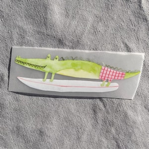 Può includere: Illustrazione ad acquerello di un alligatore verde con pantaloncini a quadri rossi, che fa surf su una tavola da surf bianca con una striscia rossa. L'alligatore sorride, su sfondo grigio.