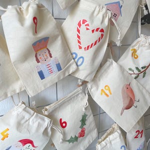 May include: A collection of off-white cotton drawstring bags featuring various holiday-themed illustrations and numbers. Designs include a nutcracker, a candy cane heart, a robin, holly berries, and a gnome, with numbers printed in red, blue, and orange.