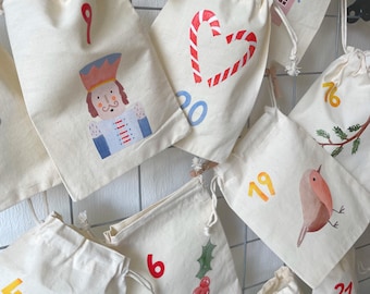 May include: A collection of off-white cotton drawstring bags featuring various holiday-themed illustrations and numbers. Designs include a nutcracker, a candy cane heart, a robin, holly berries, and a gnome, with numbers printed in red, blue, and orange.