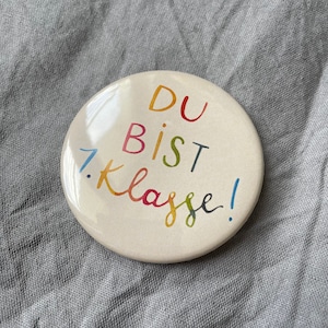 Könnte beinhalten: Ein weißer runder Knopf mit dem Text "DU BIST 1. KLASSE!" in Regenbogenfarben.
