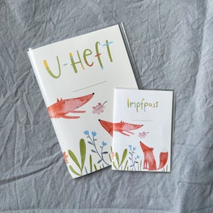 May include: Two white notebooks with a watercolor fox design. The larger notebook has the text "U-Heft" in colorful letters. The smaller notebook has the text "Impfpass" in colorful letters. The design includes foxes, leaves, and flowers.
