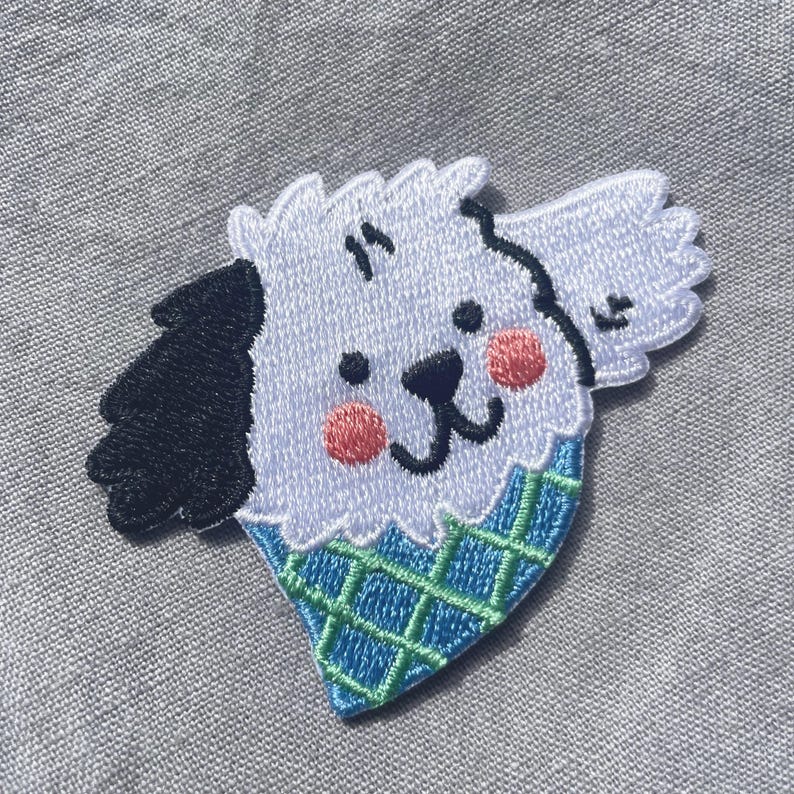 May include: An embroidered patch featuring a white dog head with one black ear, pink cheeks, and a black nose. The dog is positioned inside a blue ice cream cone shape with a green grid pattern. The embroidery has a white border and textured finish.