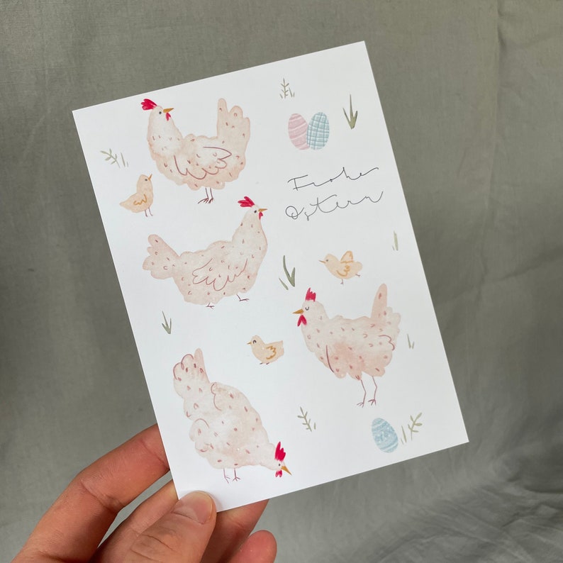 Pu&ograve; includere: Un biglietto bianco con un'illustrazione ad acquerello rosa e bianca di galline e uova. Il biglietto dice "Frohe Ostern", che in tedesco significa "Buona Pasqua".