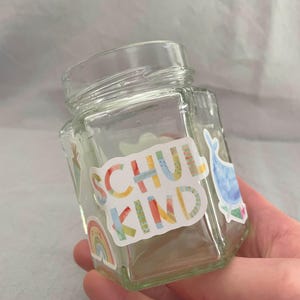Pu&ograve; includere: Un barattolo di vetro trasparente, esagonale, con coperchio a vite. Il barattolo &egrave; decorato con adesivi colorati, tra cui un arcobaleno e la parola "SCHUL KIND" in vari colori. &Egrave; visibile anche un adesivo a forma di balena.