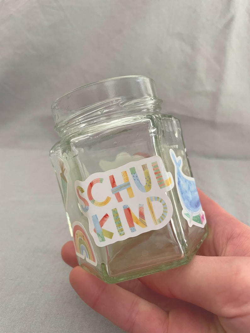 Pu&ograve; includere: Un barattolo di vetro trasparente, esagonale, con coperchio a vite. Il barattolo &egrave; decorato con adesivi colorati, tra cui un arcobaleno e la parola "SCHUL KIND" in vari colori. &Egrave; visibile anche un adesivo a forma di balena.