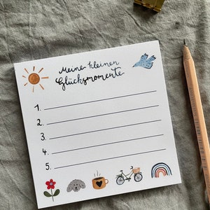 Op de afbeelding: Een wit notitieblok met de tekst "Meine kleinen Gl&uuml;cksmomente" en genummerde lijnen. Het notitieblok bevat illustraties van een zon, vogel, bloem, hond, koffiekopje, fiets en regenboog. Een potlood en een gouden paperclip zijn ook aanwezig.