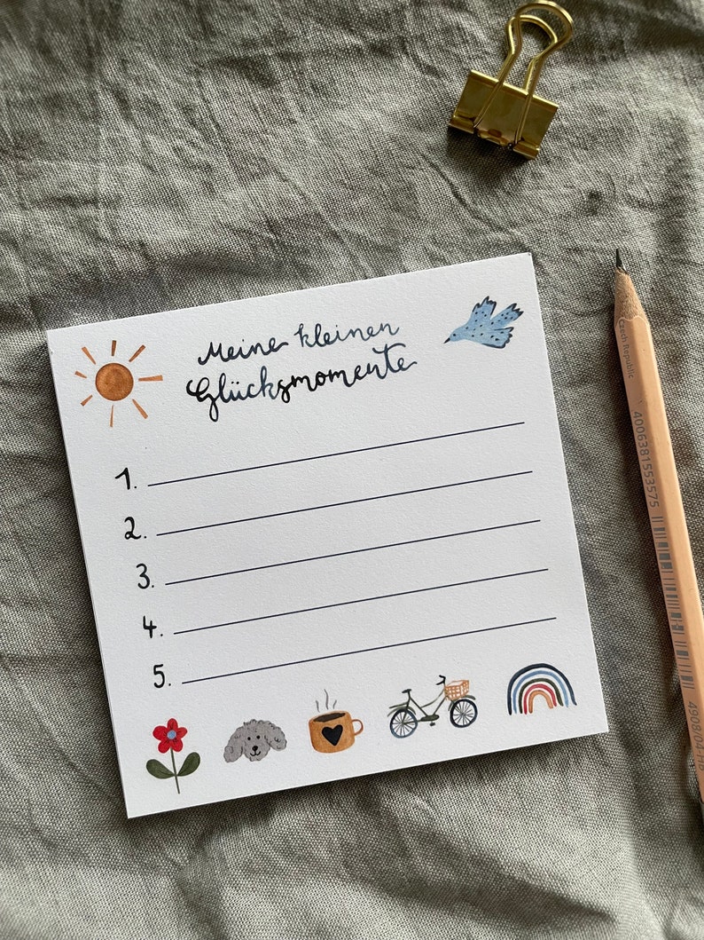 Op de afbeelding: Een wit notitieblok met de tekst "Meine kleinen Gl&uuml;cksmomente" en genummerde lijnen. Het notitieblok bevat illustraties van een zon, vogel, bloem, hond, koffiekopje, fiets en regenboog. Een potlood en een gouden paperclip zijn ook aanwezig.