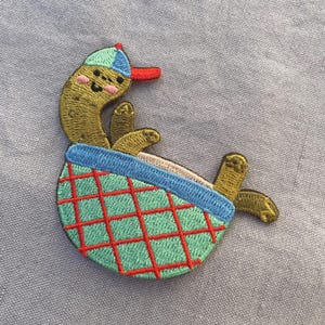 May include: Embroidered patch of a cartoon pickle wearing a baseball cap and lounging in a green and red checkered basket. The pickle has a smiling face and is in a relaxed pose. The patch is on a light blue background.