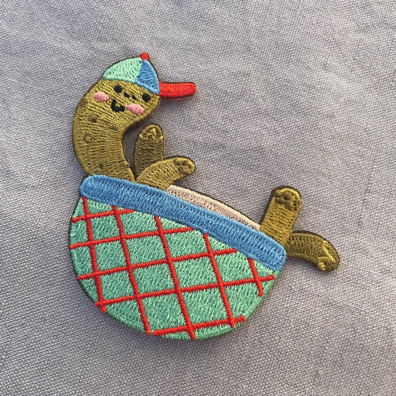May include: Embroidered patch of a cartoon pickle wearing a baseball cap and lounging in a green and red checkered basket. The pickle has a smiling face and is in a relaxed pose. The patch is on a light blue background.