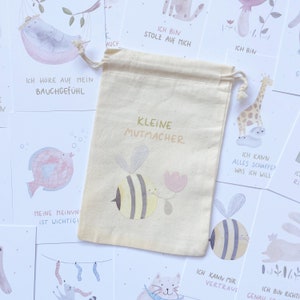 Op de afbeelding: Een set kleine aanmoedigingskaartjes met een beige trekkoordtas. De tas heeft een bij- en bloemetjesontwerp en de tekst "Kleine Mutmacher". De kaartjes tonen verschillende illustraties en positieve affirmaties in het Duits.