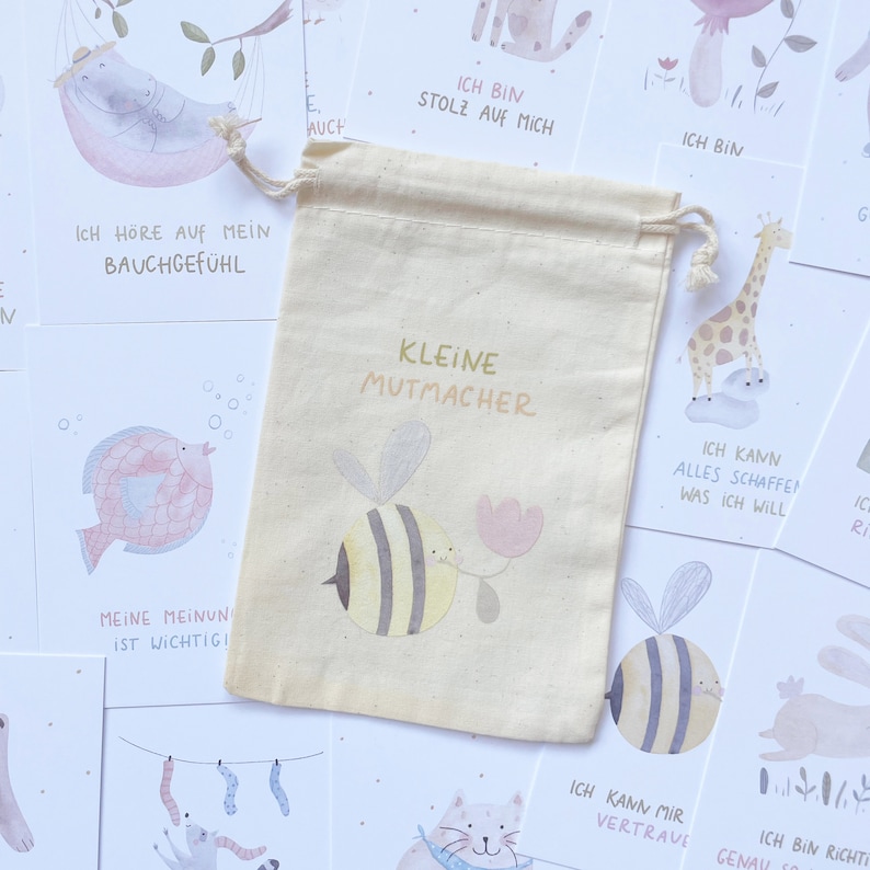 Op de afbeelding: Een set kleine aanmoedigingskaartjes met een beige trekkoordtas. De tas heeft een bij- en bloemetjesontwerp en de tekst "Kleine Mutmacher". De kaartjes tonen verschillende illustraties en positieve affirmaties in het Duits.