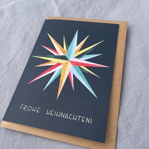 May include: A greeting card with a navy blue background and a colorful starburst design in shades of red, yellow, blue, and green. The text "FROHE WEIHNACHTEN!" is printed in white. The card is placed inside a brown envelope.