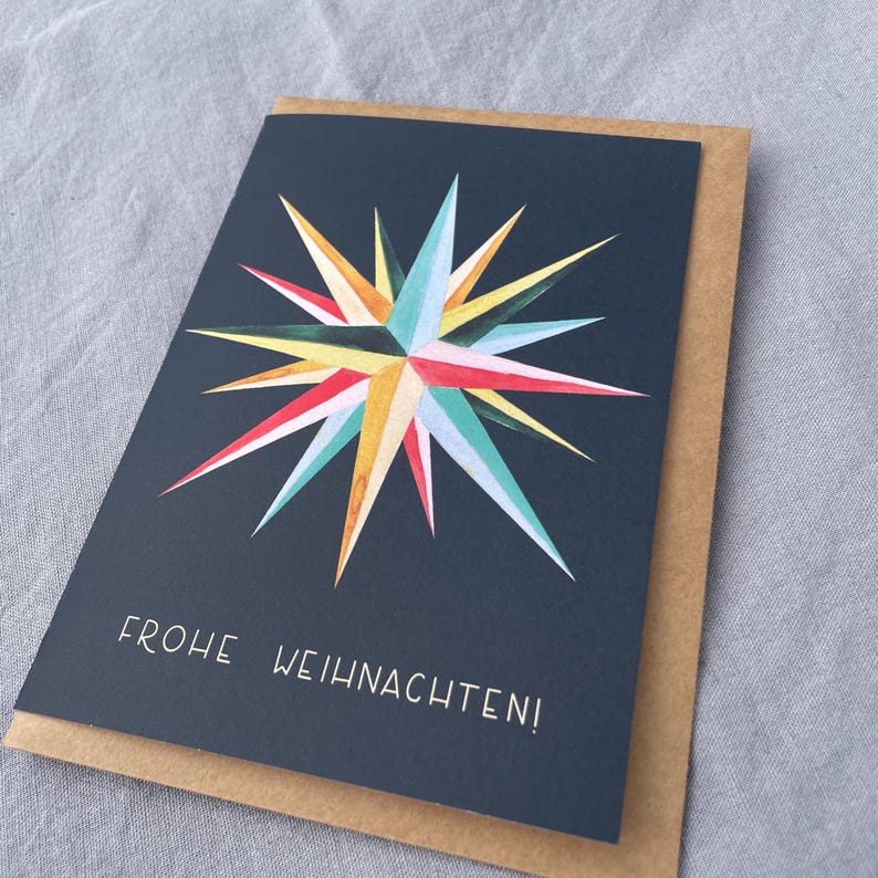 May include: A greeting card with a navy blue background and a colorful starburst design in shades of red, yellow, blue, and green. The text "FROHE WEIHNACHTEN!" is printed in white. The card is placed inside a brown envelope.
