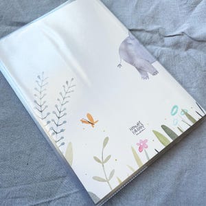 May include: A white notebook with a watercolour illustration of an elephant, butterfly, and flowers. The notebook cover has the text "Hannah Firlemann" in cursive.