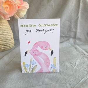 May include: A wedding greeting card with the German text "HERZLICHEN GL&Uuml;CKWUNSCH zur Hochzeit!" in green and black script. The card features two pink flamingo illustrations, a red heart, and blue flowers.