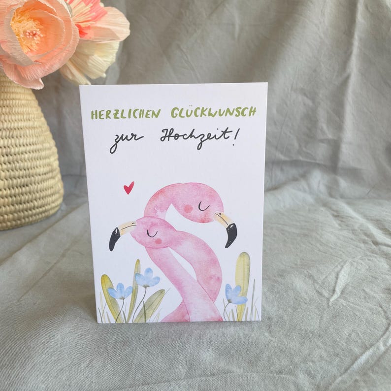 May include: A wedding greeting card with the German text "HERZLICHEN GL&Uuml;CKWUNSCH zur Hochzeit!" in green and black script. The card features two pink flamingo illustrations, a red heart, and blue flowers.