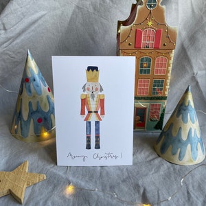 May include: A watercolor illustration of a nutcracker soldier with a red and gold uniform, a black hat, and a white beard. The illustration is on a white card with the text "Merry Christmas!" written in black ink.
