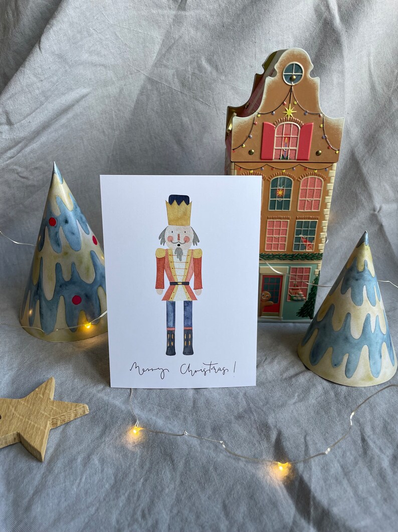May include: A watercolor illustration of a nutcracker soldier with a red and gold uniform, a black hat, and a white beard. The illustration is on a white card with the text "Merry Christmas!" written in black ink.